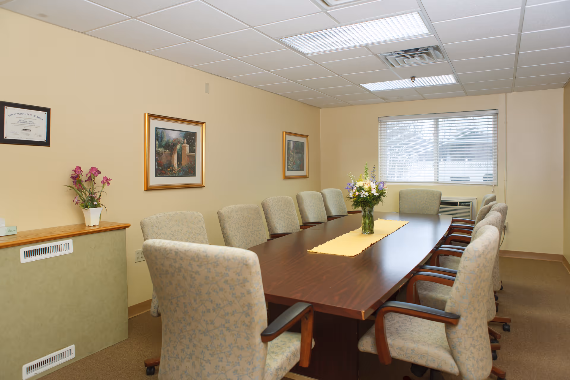 A conference room with a long wooden table surrounded by ten upholstered chairs. The table has a yellow runner and a vase with flowers in the center. The room has beige walls with two framed pictures and a certificate on one wall. There is a window with blinds and an air conditioning unit beneath it. A small shelf holds a potted plant and a box of tissues.