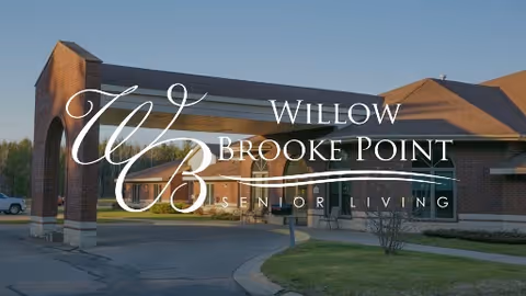Exterior view of Willow Brooke Point Senior Living facility showing a brick building with a covered entrance and a driveway under a clear blue sky.