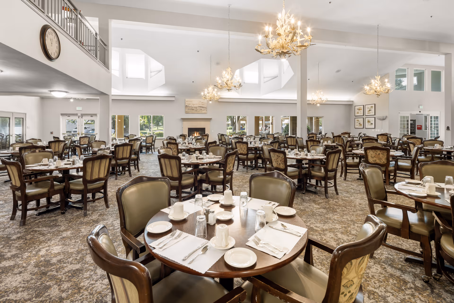 A spacious dining room in a senior living community with multiple round tables set with white plates, cups, glasses, and silverware. The room features elegant chandeliers, large windows allowing natural light, a fireplace, and comfortable upholstered chairs around each table.