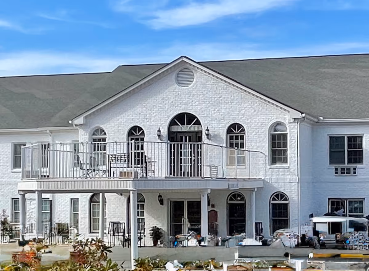 A white two-story senior living facility with a front-facing balcony and outdoor seating on the ground level.