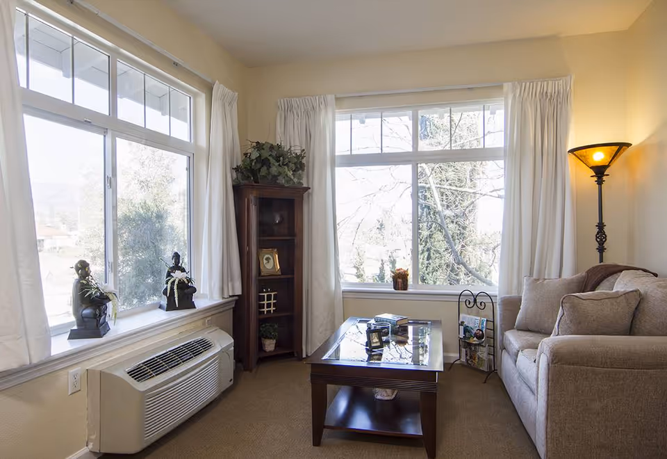 A cozy living room with large windows letting in natural light. The room features a beige sofa with cushions, a wooden coffee table with glass top, a tall wooden corner shelf with decorative items, a floor lamp with a warm light, and two small statues on the windowsill. White curtains frame the windows, and an air conditioning unit is installed below one window.