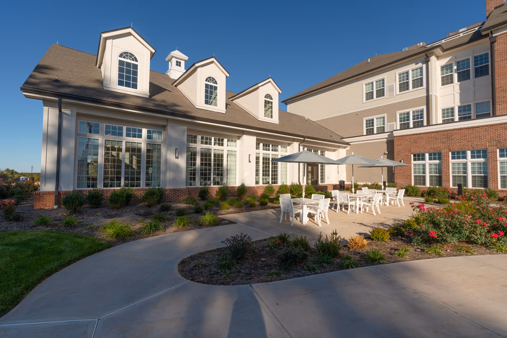 Outdoor patio area of The Culpeper facility with white tables and chairs under umbrellas, surrounded by landscaped garden beds with flowers and shrubs, adjacent to a building with large windows and dormer windows on the roof under a clear blue sky.