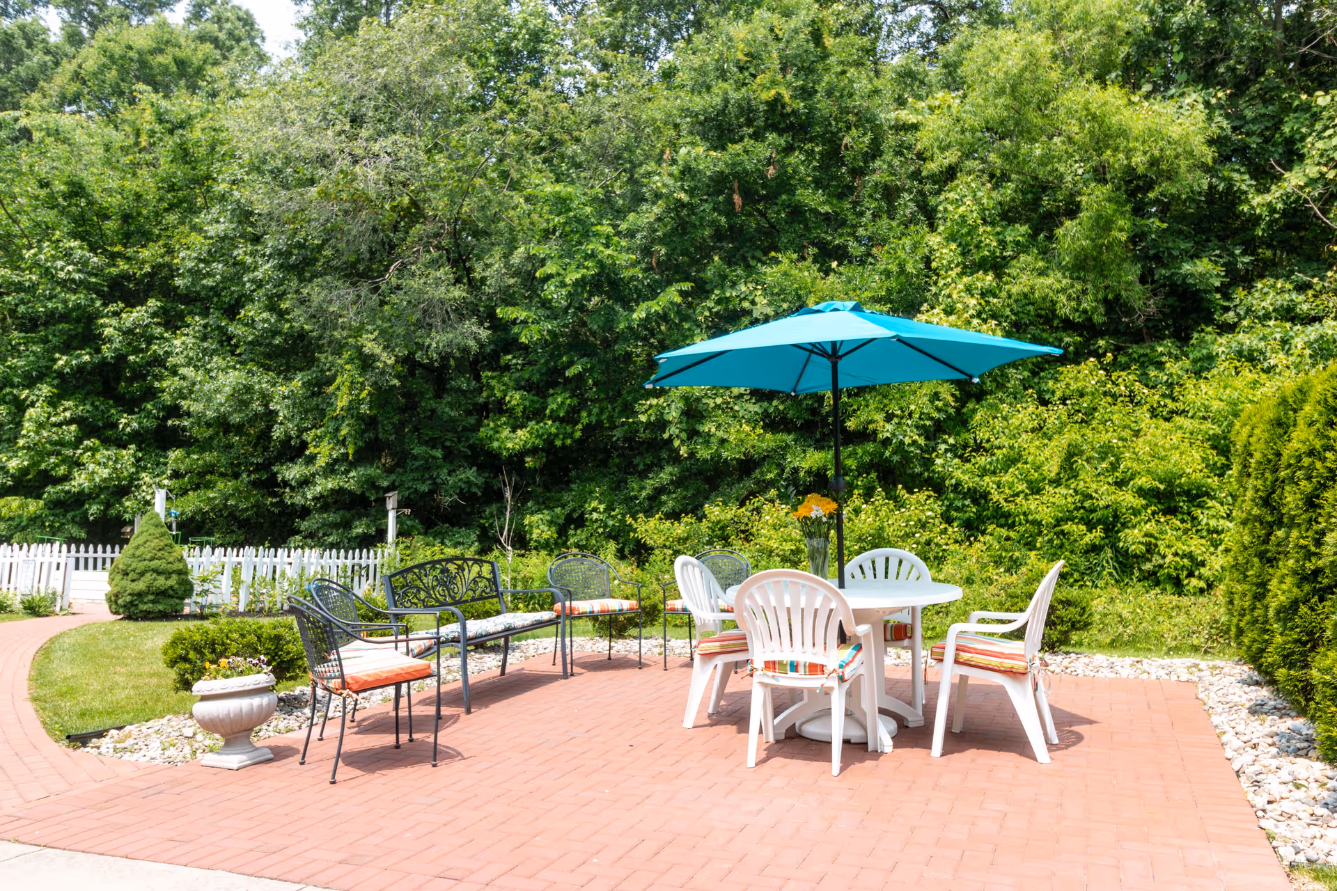 Outdoor patio area with a round white table surrounded by four white plastic chairs with striped cushions, a blue umbrella providing shade, and two black metal benches with cushions. The patio is paved with red bricks and surrounded by greenery and trees, with a white picket fence in the background.