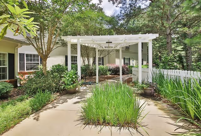 Outdoor garden area at Oaks at Fayetteville featuring a white pergola over a paved walkway surrounded by green plants, shrubs, and trees, with a white picket fence on the right side.