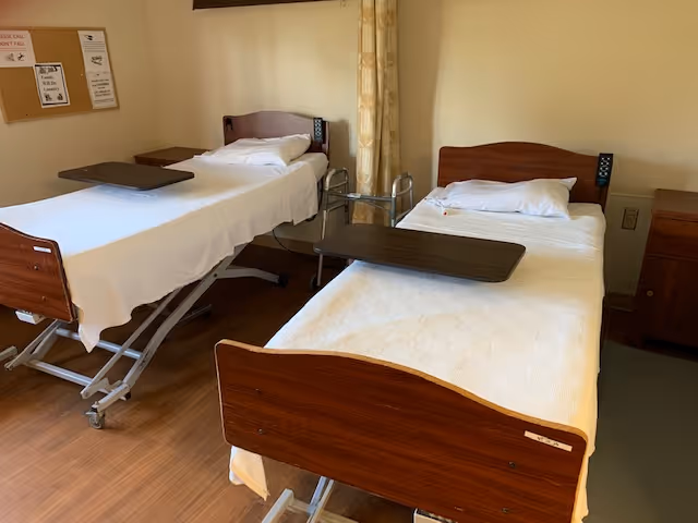 A room with two hospital-style beds, each with white bedding and a pillow. Both beds have overbed tables positioned on top. There is a small wooden nightstand between the beds and a bulletin board on the wall in the background. The floor is wooden, and the walls are painted a light beige color.