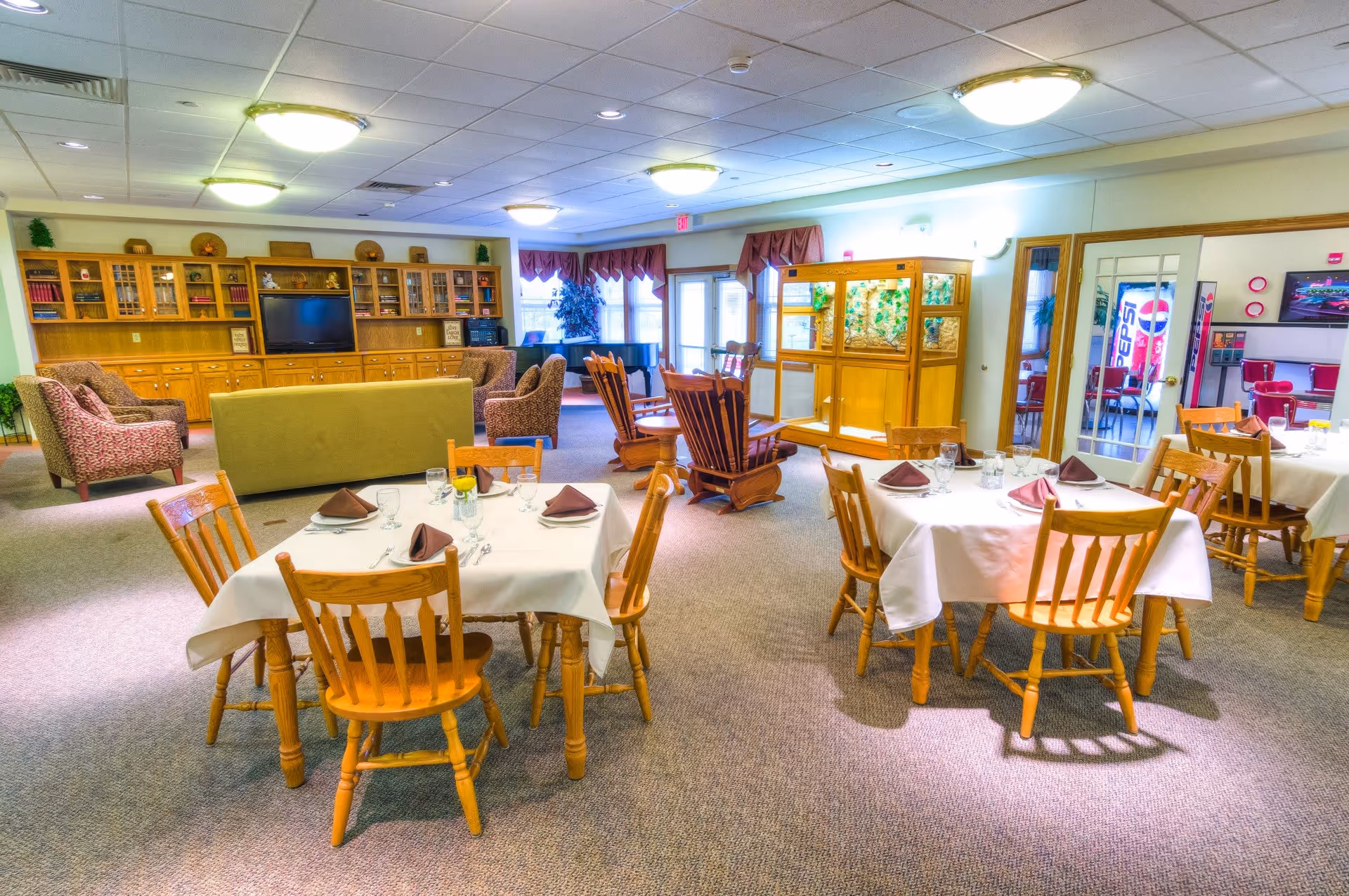 A spacious dining and lounge area in a senior living facility with several wooden tables covered with white tablecloths, set with glasses, napkins, and cutlery. The room also features comfortable upholstered chairs, a green sofa, wooden rocking chairs, a large wooden cabinet with a TV, and windows with red valances letting in natural light.