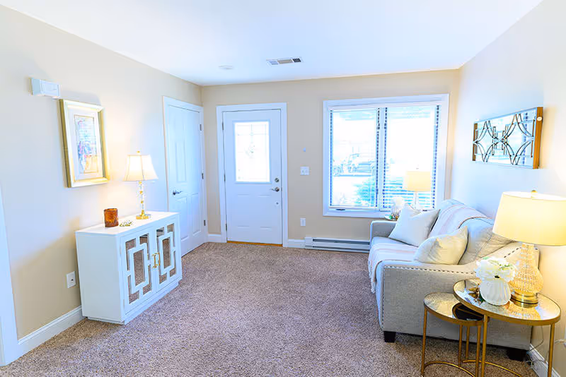 A bright living room with beige walls and carpeted floor. There is a white door with a window and a window with blinds letting in natural light. A gray sofa with cushions is placed against the wall, next to a round side table with a lamp and decorative items. On the opposite wall, there is a white cabinet with a lamp and framed artwork above it. A decorative wall piece hangs above the sofa.
