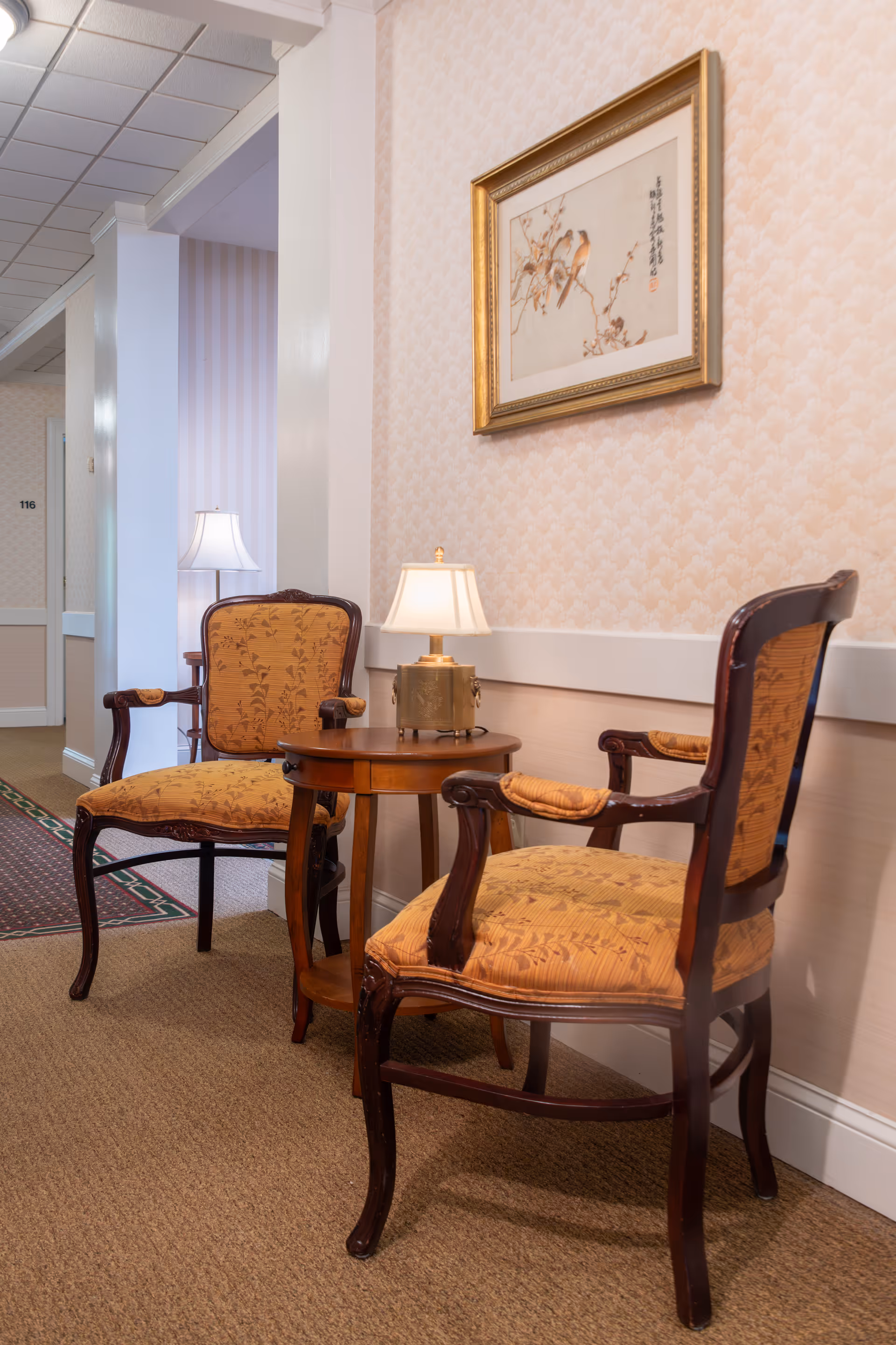 Two vintage-style upholstered armchairs with wooden frames are positioned around a small round wooden table with a lamp on it, set against a wallpapered wall with a framed painting of birds on a branch. The setting appears to be a quiet hallway in an assisted living facility, with a carpeted floor and soft lighting.