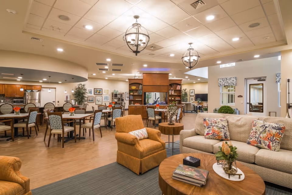 A spacious and well-lit common area in a senior living facility featuring a cozy seating area with a beige sofa and orange armchairs, a wooden coffee table with books and a flower vase, multiple dining tables with chairs, a kitchen area with stainless steel appliances, decorative plants, and framed artwork on the walls.