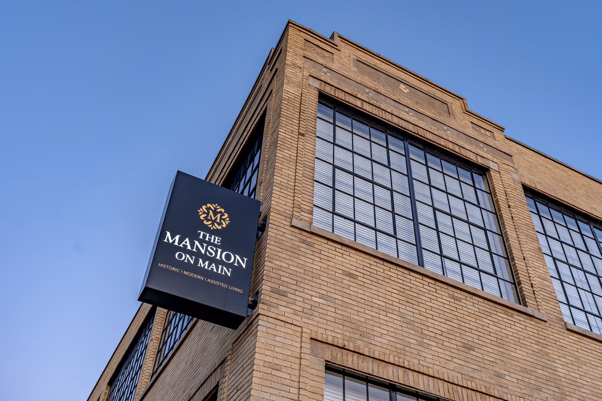 Brick exterior corner of The Mansion on Main with a black hanging sign and large industrial-style windows against a clear blue sky.