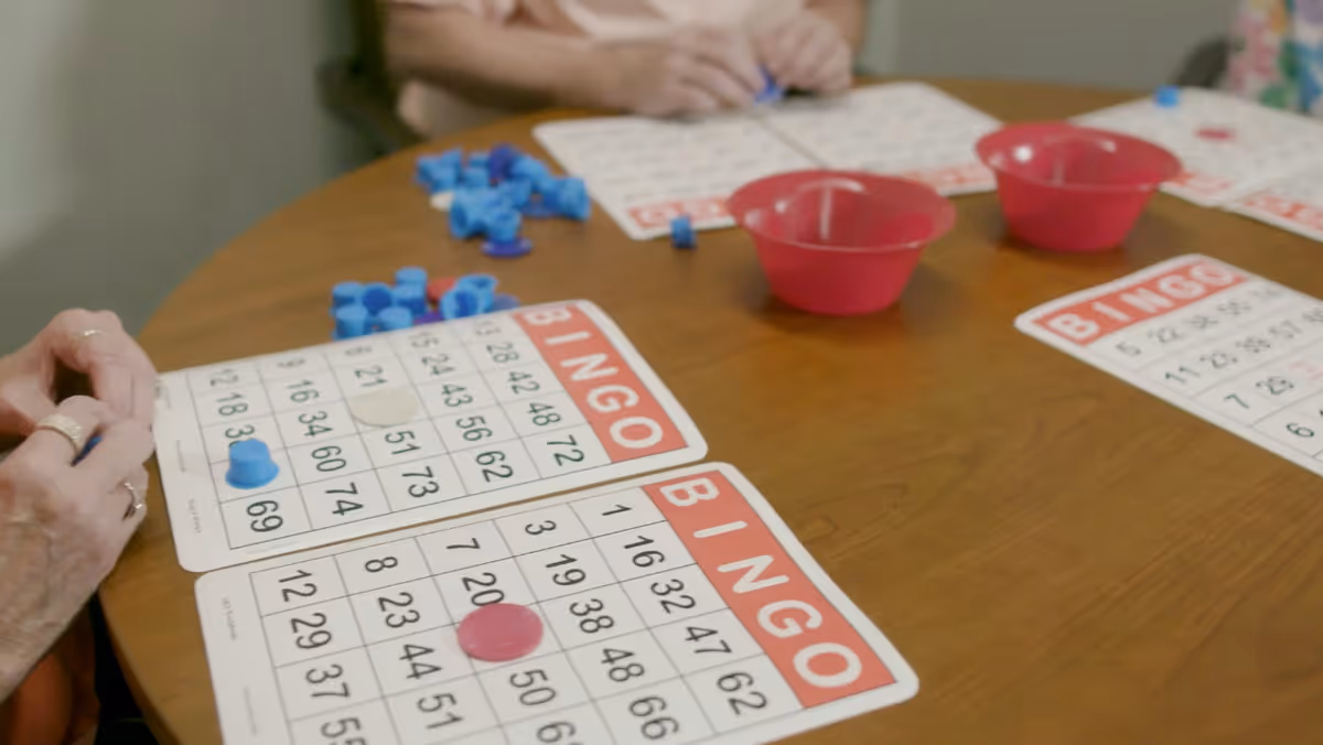 Close-up view of a wooden table with bingo cards and colorful bingo chips. Several hands are seen placing chips on the cards, indicating a game in progress. Pink bowls are also on the table holding additional bingo chips.