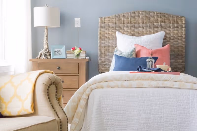 A cozy bedroom with a wicker headboard, white bedding, and colorful pillows in blue, pink, and patterned designs. A wooden nightstand beside the bed holds a lamp, a framed photo, books, and a small vase with flowers. A beige armchair with nailhead trim and a yellow patterned blanket is partially visible in the foreground.