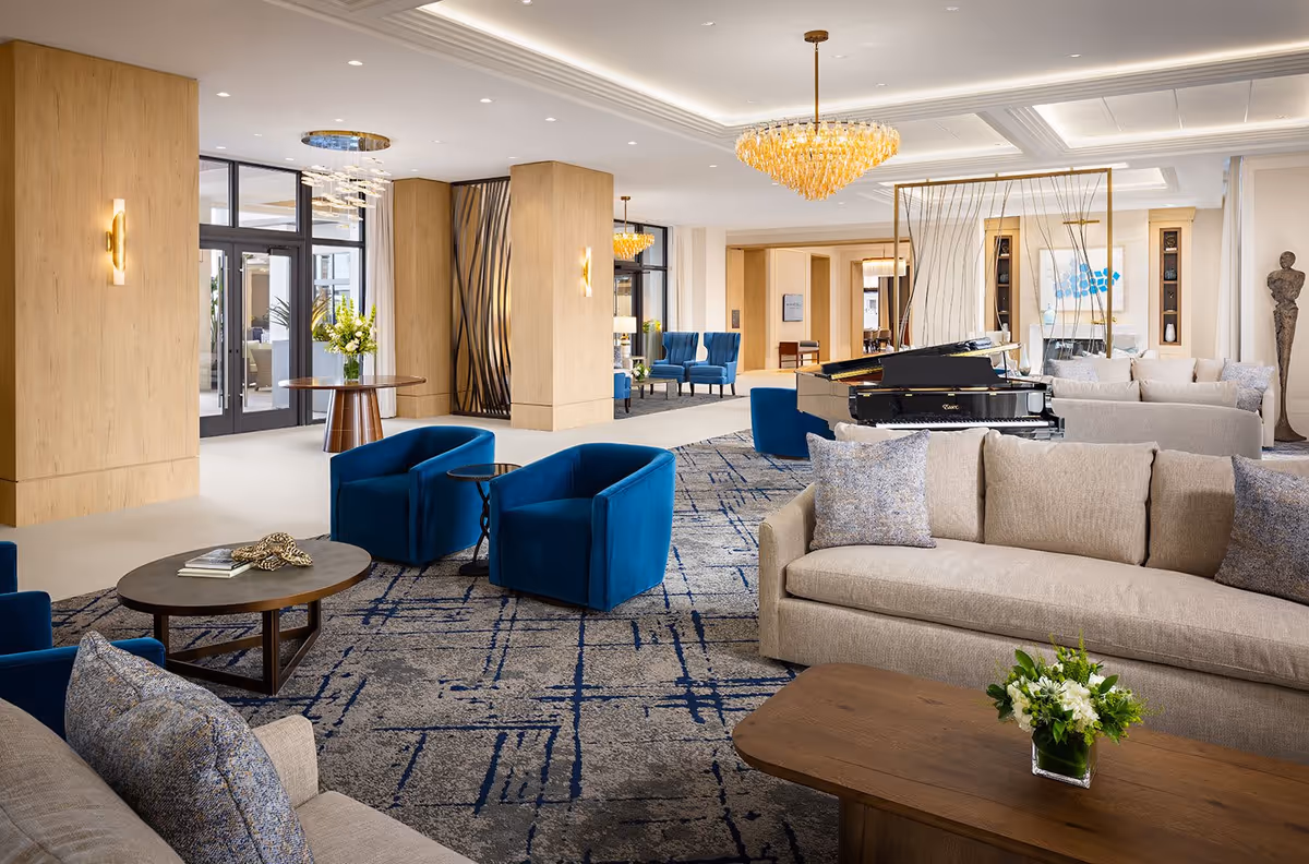 A spacious and elegant senior living facility lounge area featuring beige sofas with patterned cushions, blue velvet armchairs, a grand piano, modern chandeliers, wooden tables with floral arrangements, and large windows allowing natural light.