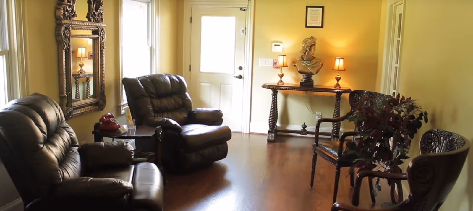 Seated living room with two leather recliners, a decorative mirror, a console table with lamps and a bust, and carved wooden chairs on a hardwood floor.