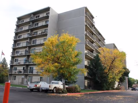 Multi-story senior living facility with balconies, parked cars, and fall-colored trees.