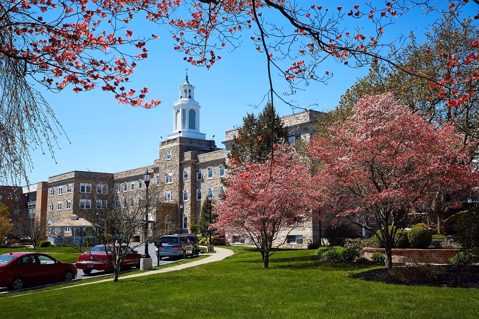 Stone-faced senior living building with a white cupola, landscaped lawn and pink flowering trees in front under a blue sky.