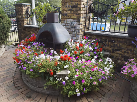 Outdoor patio area with a circular brick planter filled with colorful flowers including red, white, purple, and pink blooms surrounding a black metal outdoor fireplace. The area is enclosed by a brick wall with black wrought iron fencing and a swimming pool is visible in the background.