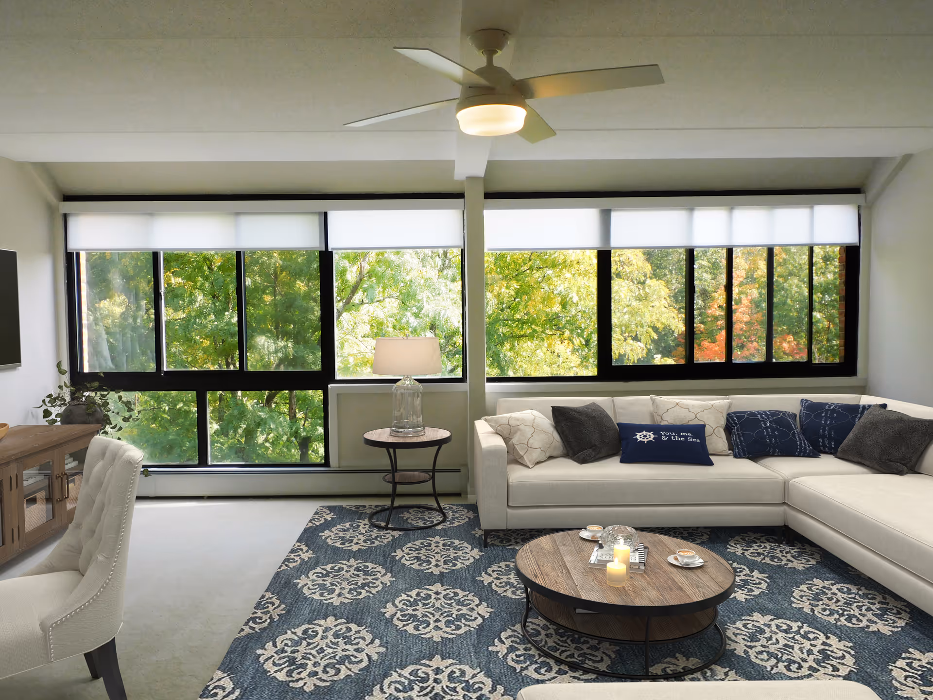 A bright living room with large windows showing green trees outside. The room features a white sectional sofa with various pillows, a round wooden coffee table with candles and cups, a side table with a lamp, a ceiling fan with light, a patterned blue and white rug, a white upholstered chair, and a wooden TV stand with a flat-screen TV mounted on the wall.