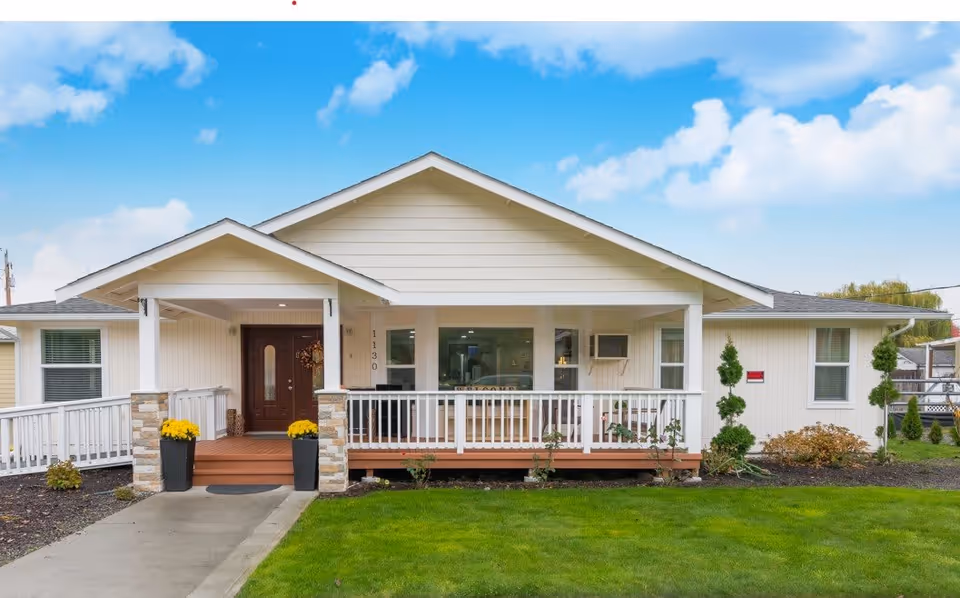 Front exterior view of a single-story adult family home with a covered porch, white railing, and a brown front door. The house is painted white with a gray roof and has a well-maintained green lawn and some small shrubs and plants around the porch. The sky is blue with scattered clouds.