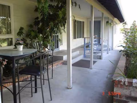 Outdoor covered walkway with tables, chairs, and potted plants beside resident rooms.