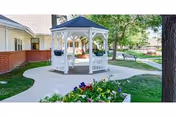 White gazebo in a landscaped courtyard with a paved walkway, benches, flowers, and adjacent buildings.