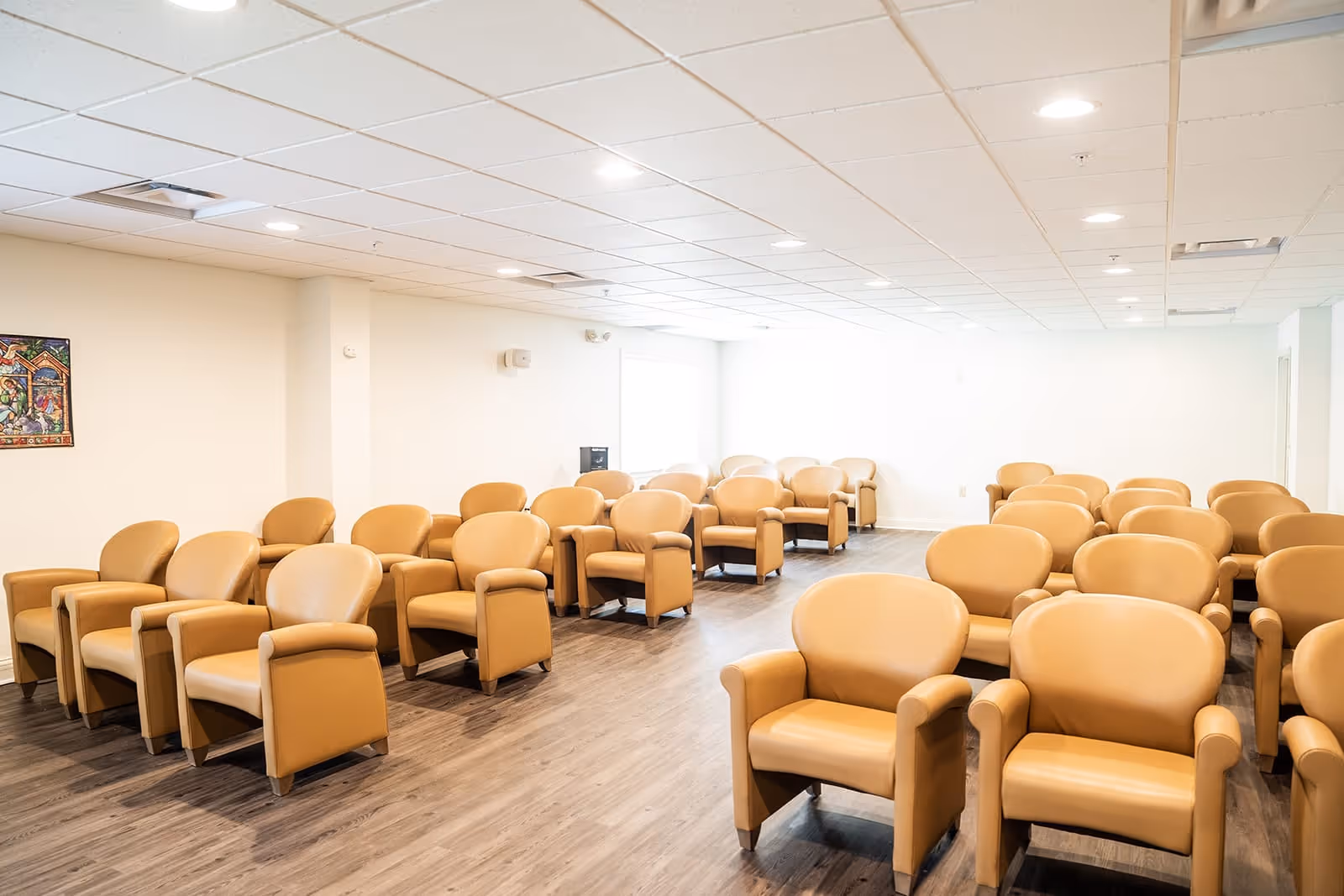 A bright room with multiple rows of tan leather armchairs arranged facing forward on a wood floor, with white walls and a drop ceiling with recessed lighting.