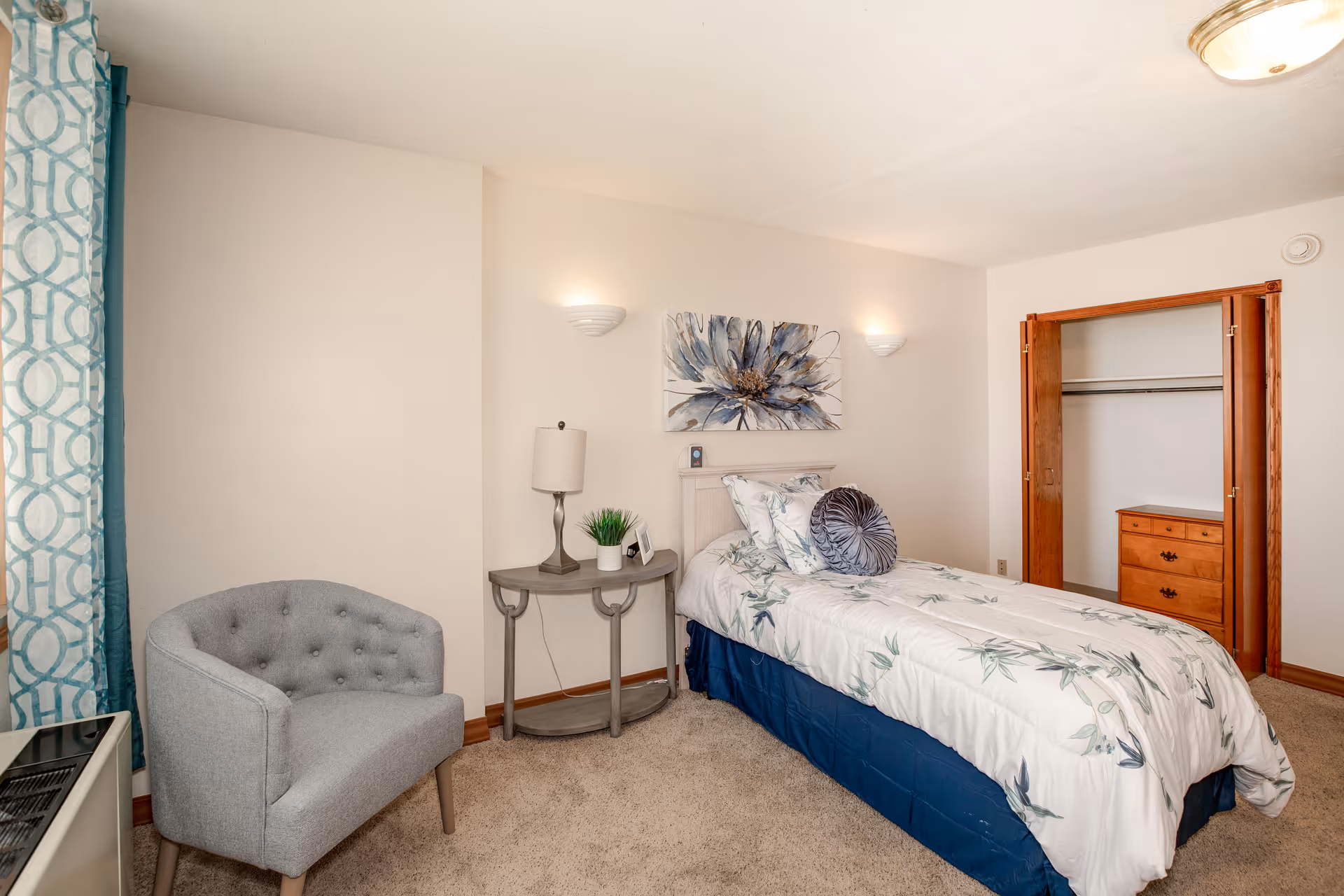 A neatly arranged bedroom with a single bed covered in white and blue floral bedding. There is a gray upholstered chair near a window with patterned blue and white curtains. A small gray side table with a lamp, a small plant, and a picture frame is next to the bed. The room has beige walls, carpeted floor, and an open closet with wooden doors and a chest of drawers inside. A large floral painting hangs above the bed.
