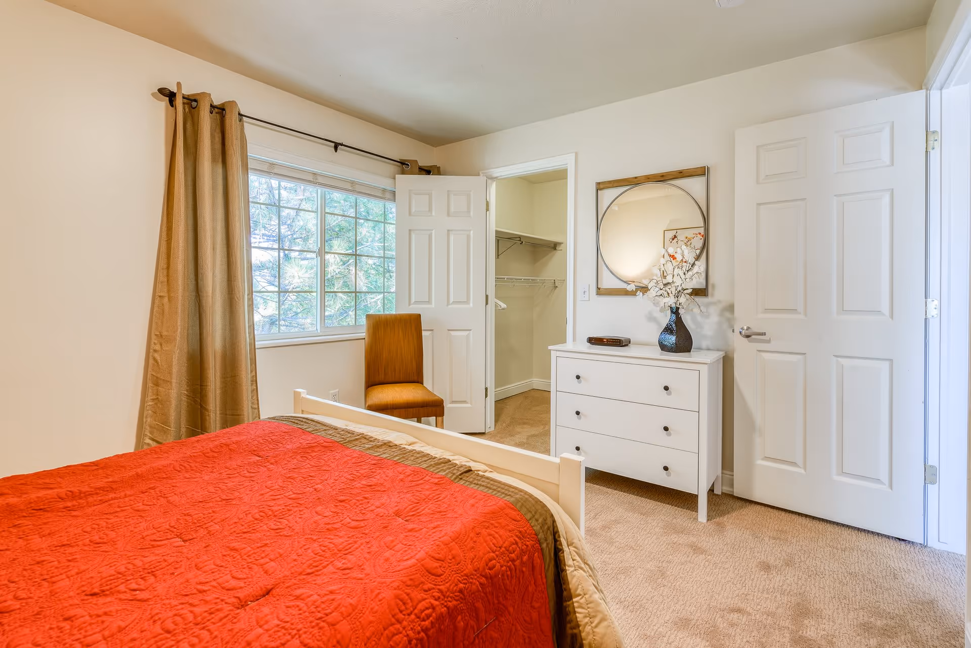 A bedroom with a bed covered in a red quilt, a window with beige curtains, a wooden chair, an open door leading to a walk-in closet, and a white dresser with a round mirror and a vase with white flowers on top.