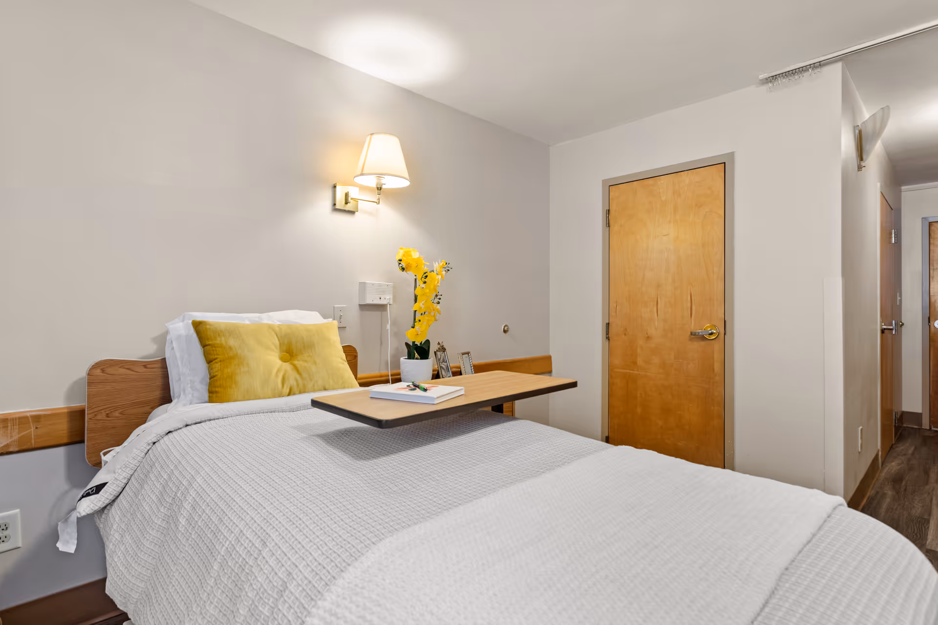 A simple bedroom in a senior living facility with a single bed covered in a white textured blanket and a yellow pillow. A wooden overbed table holds a book and a small potted plant with yellow flowers. The room has beige walls, a wooden door, and a wall-mounted lamp above the bed providing warm light.
