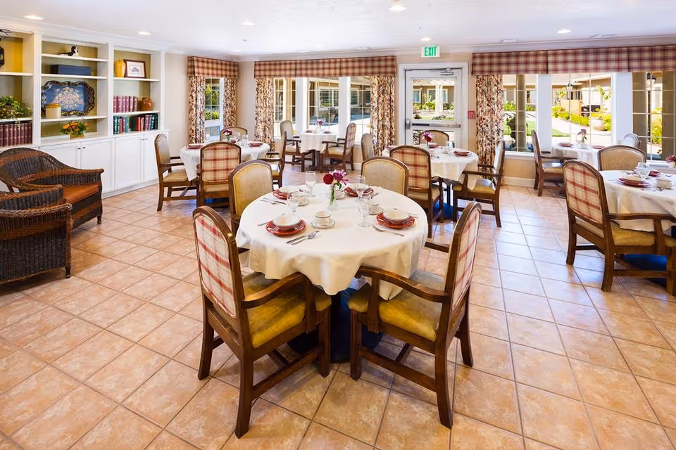 A bright and inviting dining room with several round tables covered in white tablecloths, each set with plates, cups, and utensils. The chairs have wooden frames with cushioned seats and backs, some upholstered in plaid fabric. Large windows with floral curtains allow natural light to fill the room, and built-in white shelves with books and decorative items are visible on the left side. The floor is tiled with a warm-toned pattern.