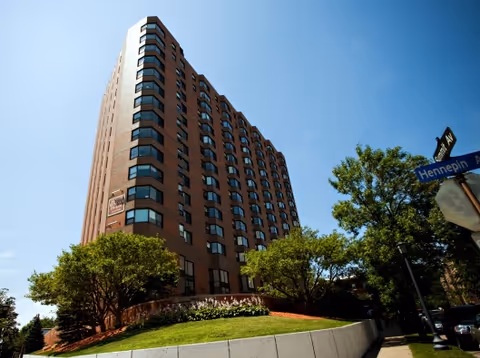 Tall multi-story brick building with many windows, surrounded by trees and a grassy area under a clear blue sky. A street sign for Hennepin Ave is visible on the right side of the image.