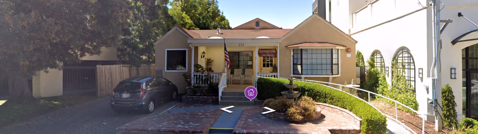 Front entrance of a small tan building with a porch, wheelchair ramp, circular fountain, and a parked car.
