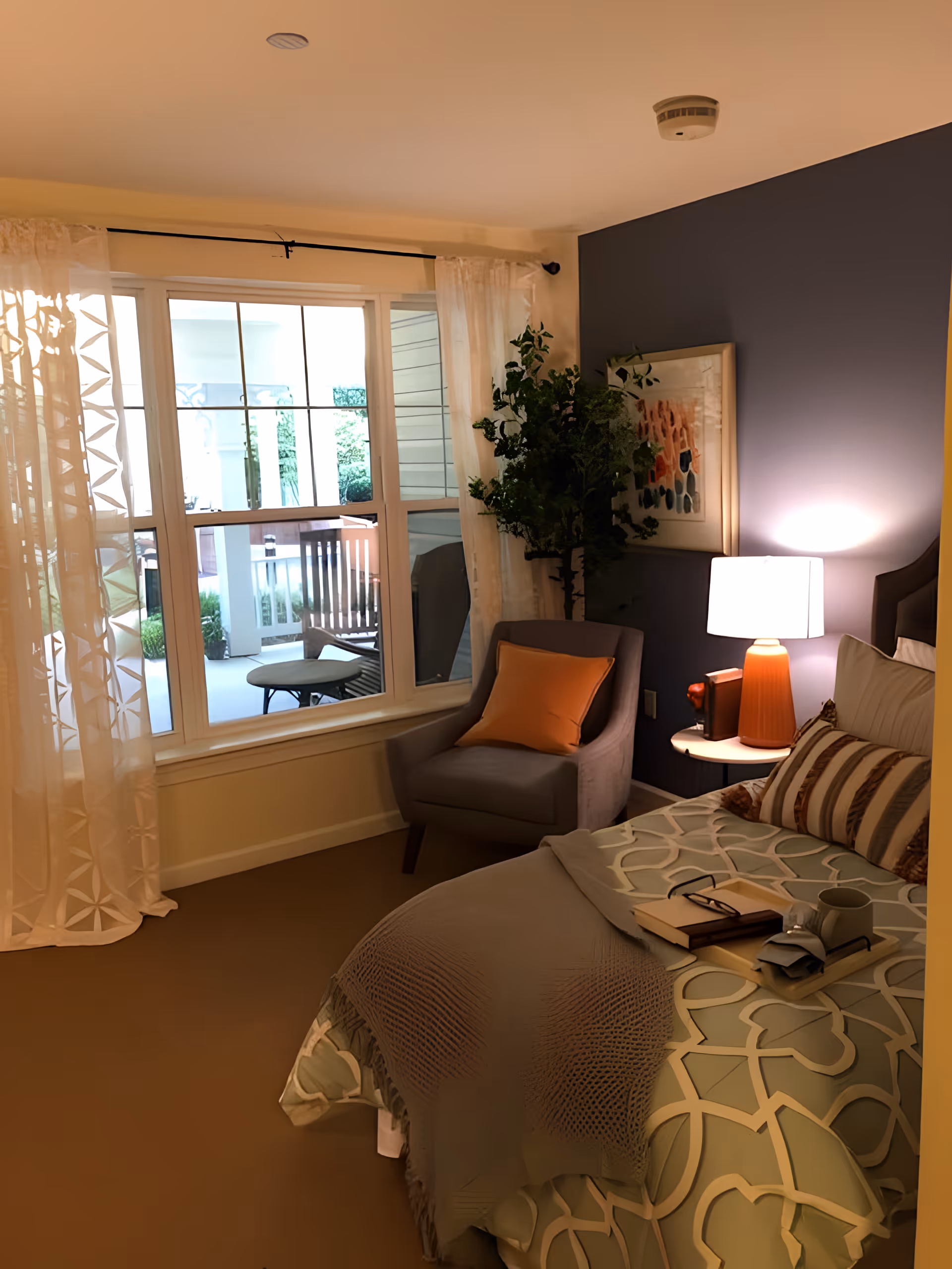 Cozy, well-lit bedroom featuring a bed with patterned bedding, an armchair by a large window overlooking a patio, and a bedside table with a lamp.
