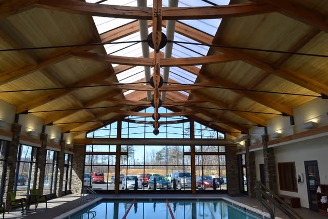 Indoor swimming pool area with large floor-to-ceiling windows showing a parking lot outside. The ceiling features wooden beams and skylights allowing natural light to fill the space. There are chairs and benches along the sides of the pool.