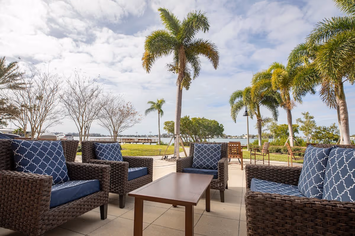 Outdoor patio area with four wicker armchairs featuring blue patterned cushions arranged around a rectangular wooden coffee table. The patio overlooks a grassy area with palm trees and a body of water in the background under a partly cloudy sky.
