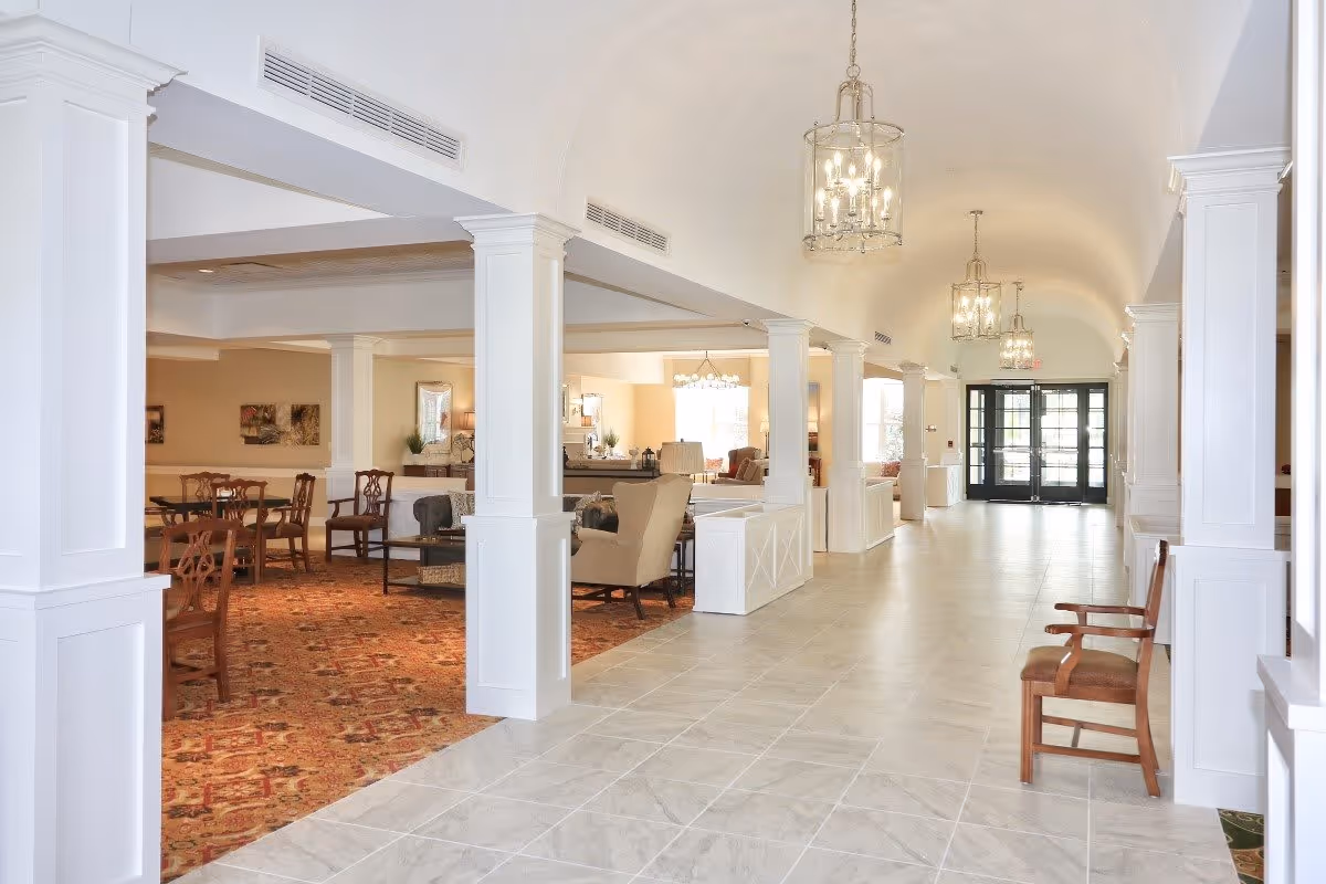 Bright and spacious senior living facility interior with a long tiled hallway featuring white columns and hanging chandeliers. To the left, there is a carpeted area with wooden chairs and tables, and a cozy seating area with armchairs and a coffee table. The far end shows large glass doors allowing natural light to enter.