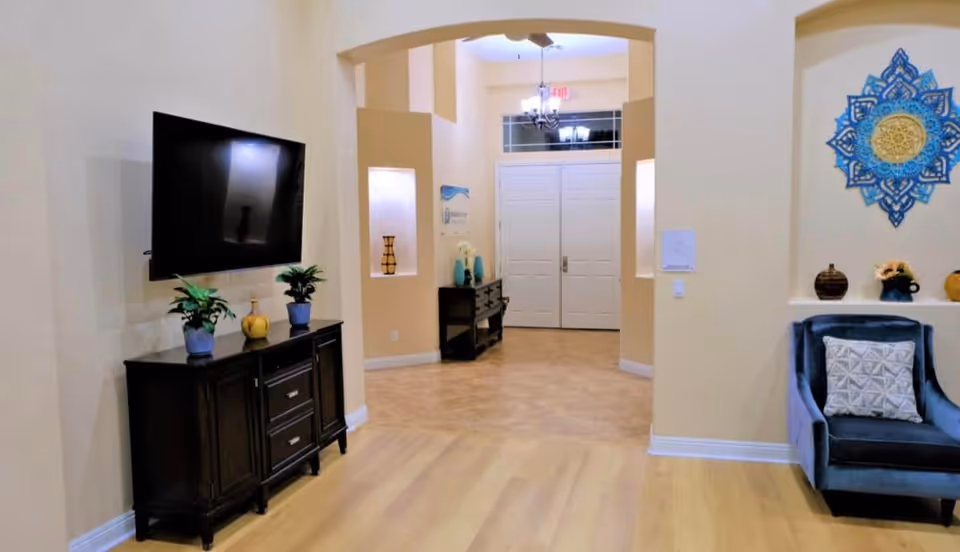 Interior view of a living area in Bethesda Care Home featuring a mounted flat-screen TV above a dark wooden cabinet with two potted plants and a decorative item. To the right, there is a blue armchair with a patterned cushion and decorative wall art above it. The room opens into a hallway with double white doors and additional furniture and decor visible.