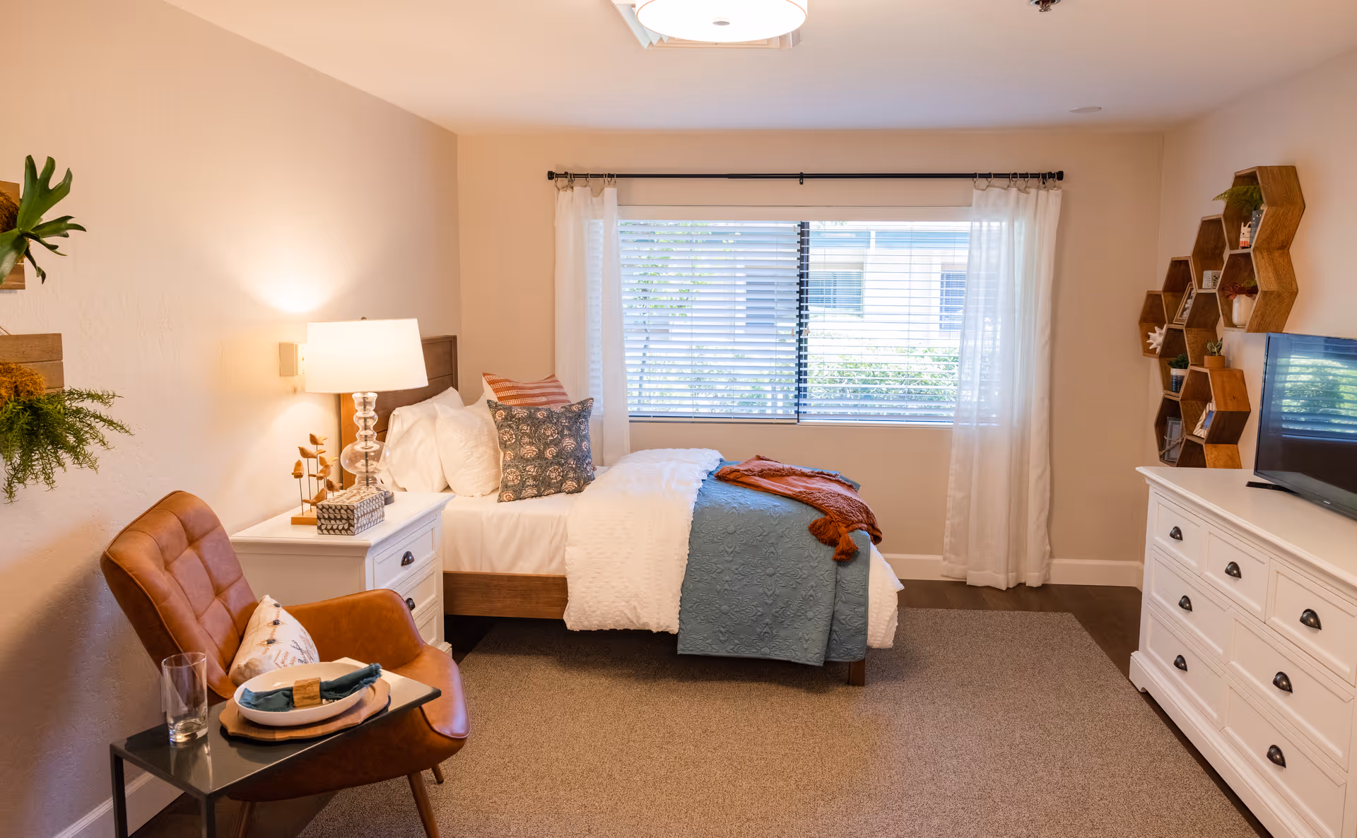 Well-lit furnished bedroom with a single bed, bedside table and lamp, armchair, dresser with TV, and a window with blinds.