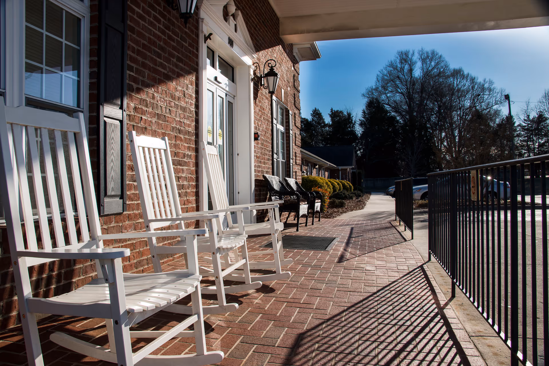 A brick building exterior with a covered porch featuring white rocking chairs and black chairs. The porch overlooks a paved walkway and parking area with trees in the background under a clear blue sky.
