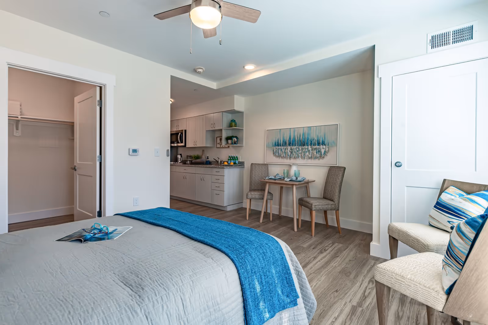 Bright studio bedroom with a bed dressed in gray and blue, a kitchenette, a small dining table with two chairs, and additional seating.