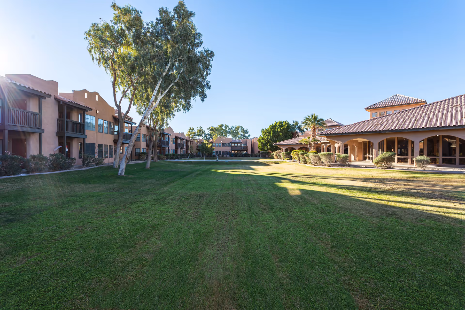 Wide view of a senior living facility's outdoor courtyard with well-maintained green grass, trees, and shrubs. The buildings surrounding the courtyard have a southwestern architectural style with stucco walls and tiled roofs. The sky is clear and blue.
