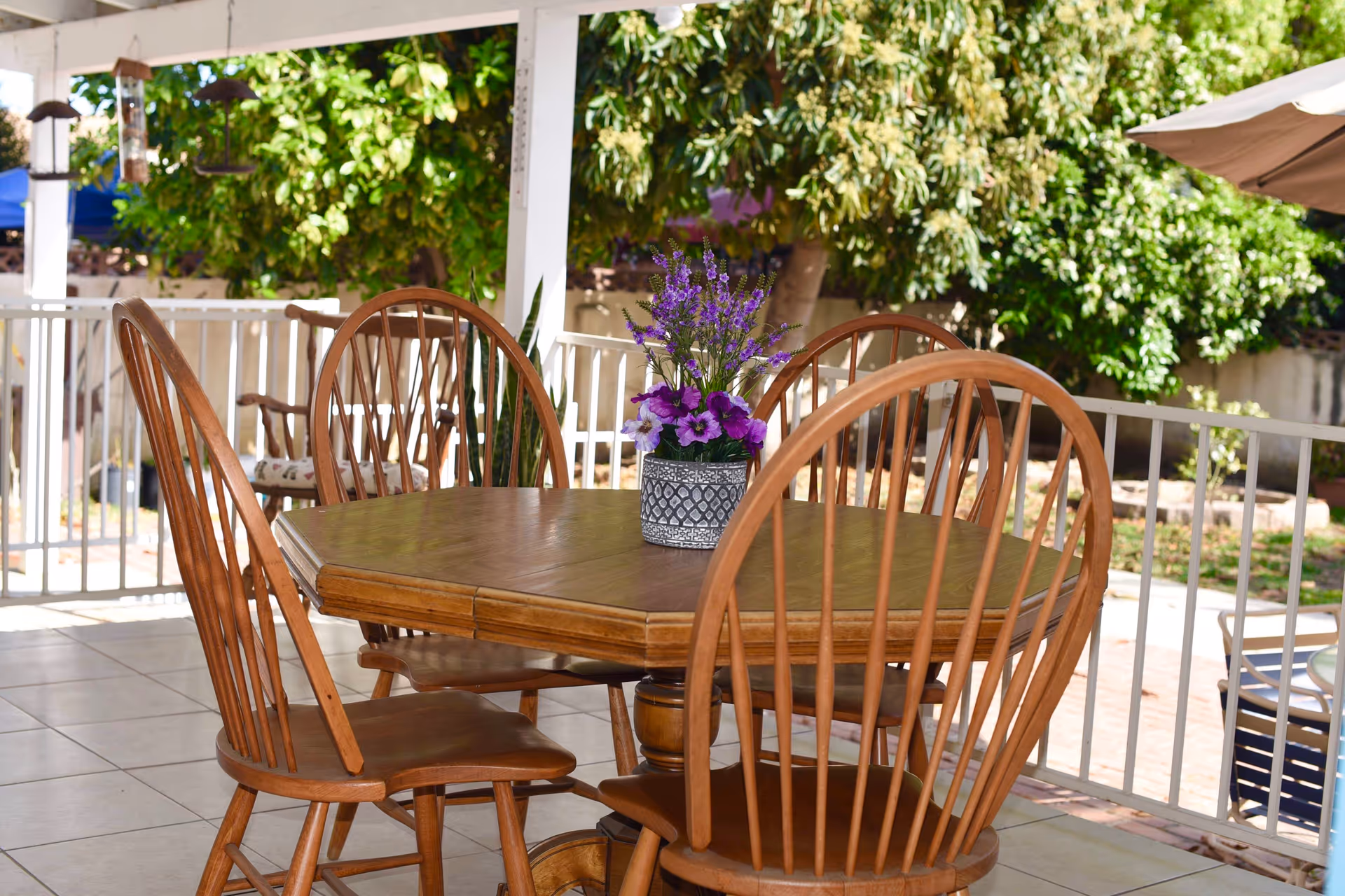 Wooden outdoor dining table with chairs and a potted purple flower centerpiece on a covered patio with trees in the background.