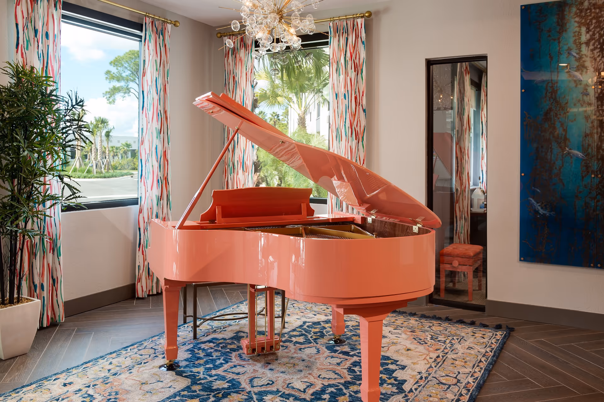 A bright coral-colored grand piano with its lid open sits on a patterned blue and beige rug in a well-lit room. The room features two large windows with colorful curtains, a tall green plant in a white pot, a modern chandelier, a narrow mirror reflecting part of the piano and a coral-colored piano bench, and a large abstract blue and brown wall art piece.