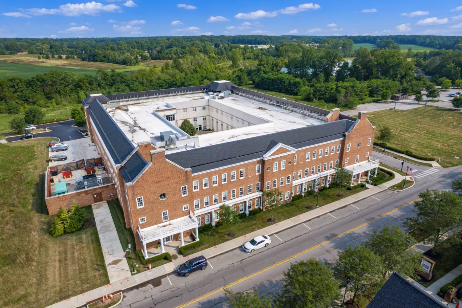 Aerial view of a large, three-story brick building with a flat white roof and a central courtyard. The building is surrounded by greenery and parking areas, with a road running alongside it. The sky is blue with scattered clouds.