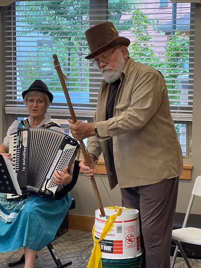 An elderly woman playing an accordion seated next to an elderly man wearing a brown top hat and beige jacket who is playing a homemade percussion instrument made from a bucket and a stick in a room with large windows and blinds.