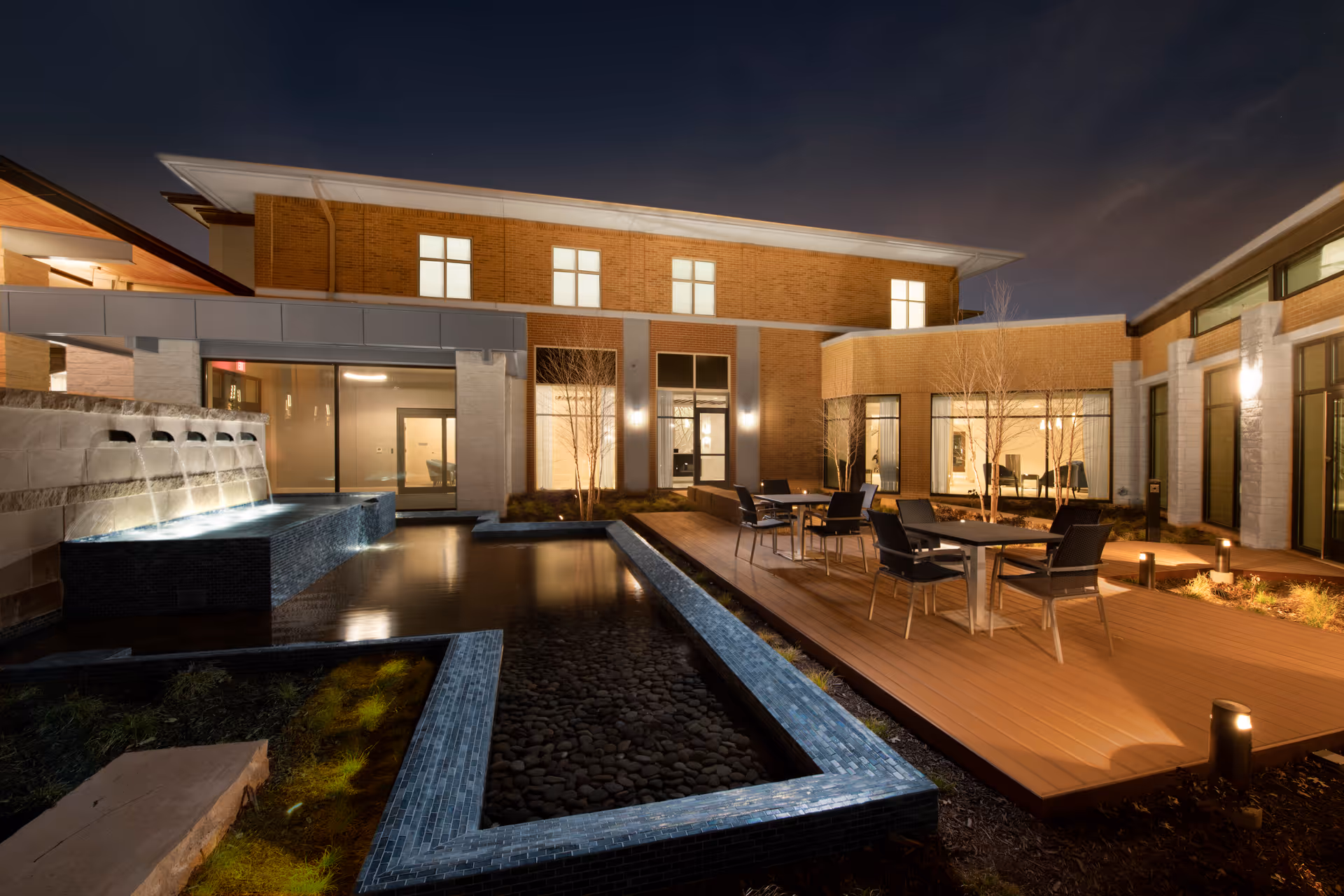 Night view of an outdoor courtyard area at Presbyterian Village North featuring a modern water fountain with cascading water into a rectangular pool, surrounded by a wooden deck with tables and chairs. The building's exterior walls are illuminated, showcasing large windows and brickwork.
