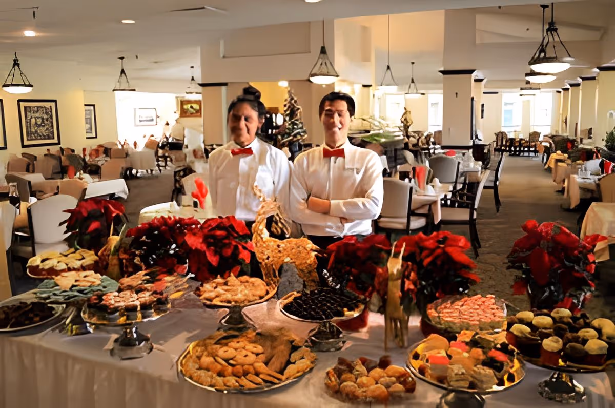 Two servers stand behind a dessert buffet with trays of pastries and poinsettias in a large dining room.