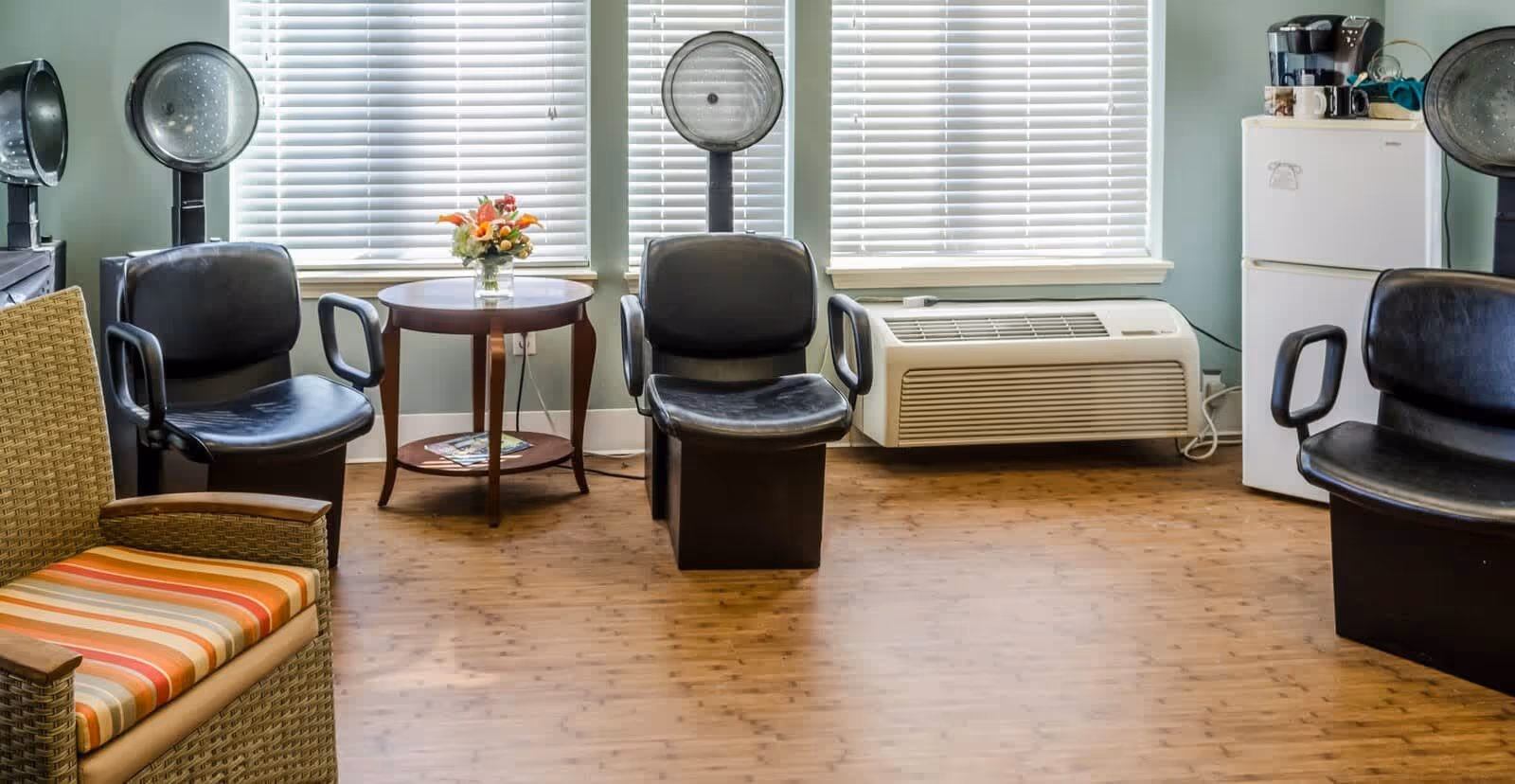 Interior of a hair salon area with three black salon chairs under hair dryers, a small round wooden table with a flower vase, a wicker chair with striped cushion, a white mini refrigerator, and a coffee maker on top. The room has wooden flooring and large windows with white blinds.