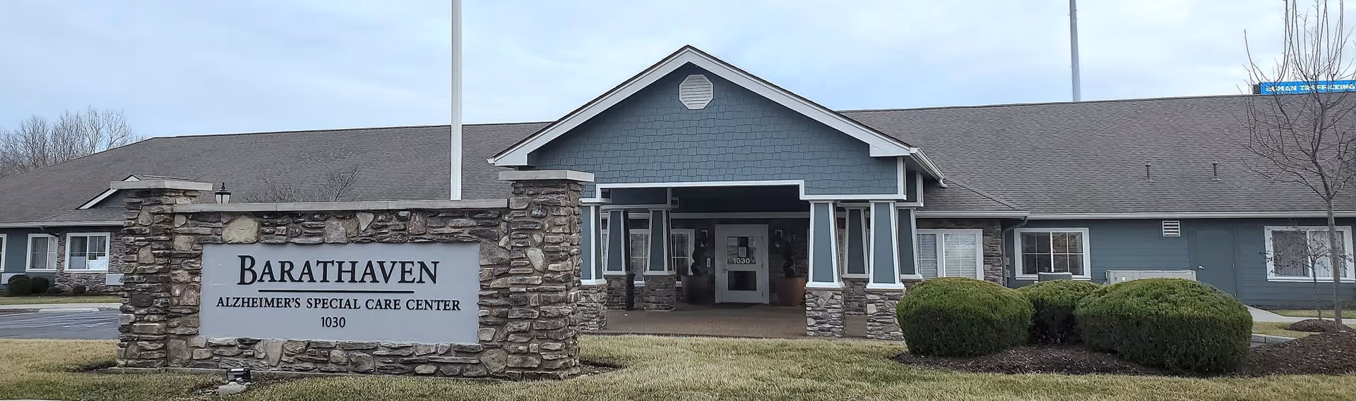 Front exterior view of Barathaven Alzheimer's Special Care Center, a single-story building with a stone sign in front displaying the facility's name and address. The building has a covered entrance with stone pillars and a gray roof, surrounded by trimmed bushes and a lawn.