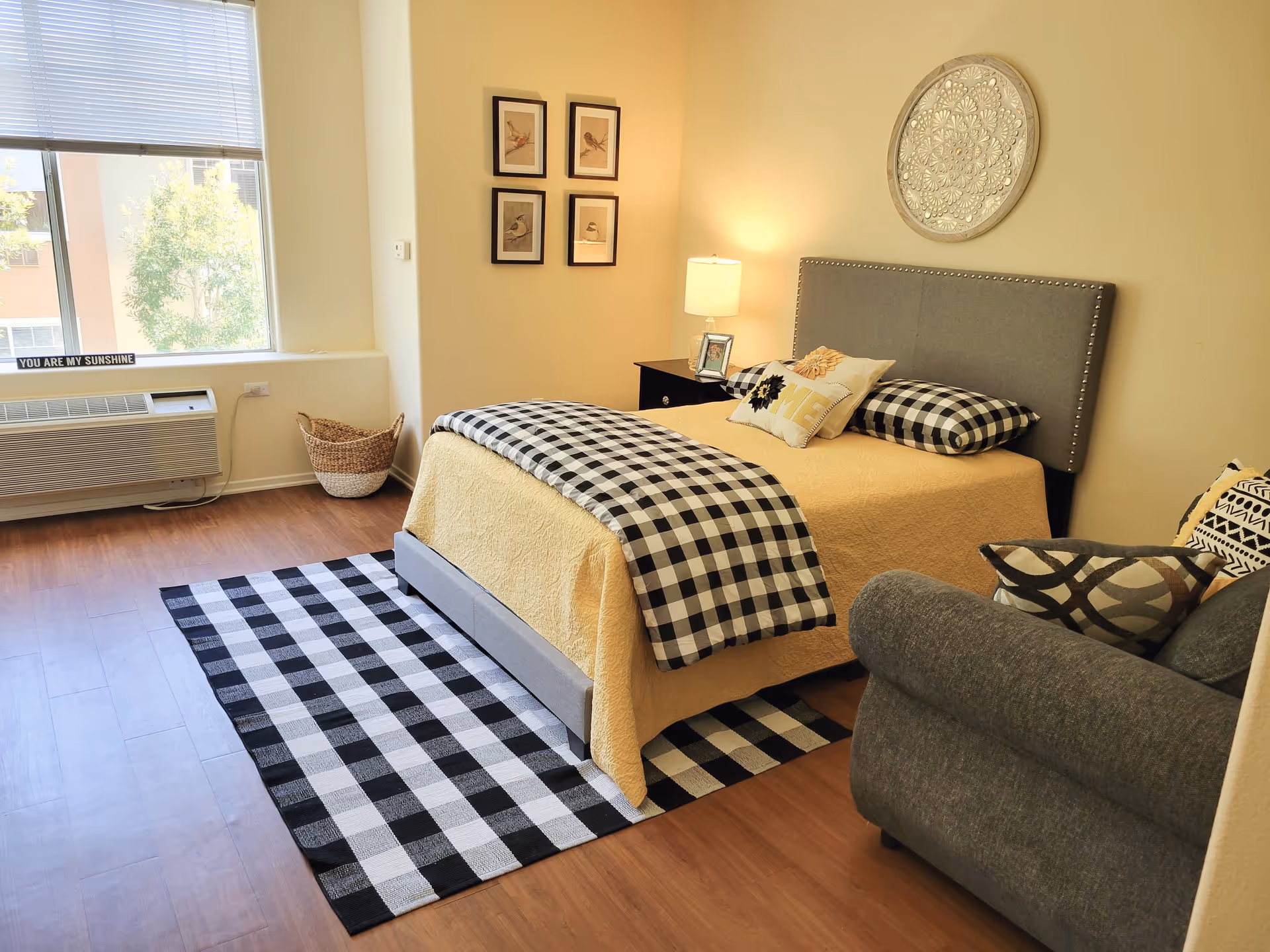 A cozy bedroom with a bed covered in a yellow quilt and black-and-white checkered blanket and pillows. There is a gray upholstered headboard with a decorative round wall piece above it. A nightstand with a lamp and picture frame is beside the bed. A gray armchair with patterned pillows is in the corner. The room has wooden flooring, a black-and-white checkered rug, a large window with blinds, and a basket near the window.
